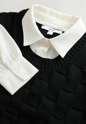 Gilet en tricot noir avec un motif à carreaux texturé, associé à une chemise blanche à col avec des poignets boutonnés. Étiquettes visibles.