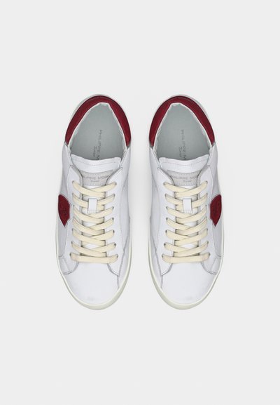 Ζευγάρι λευκών δερμάτινων αθλητικών παπουτσιών με μπεζ κορδόνια και κόκκινες λεπτομέρειες στη φτέρνα και τις πλευρές, φωτογραφημένα από πάνω σε απλό φόντο.