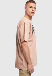 Lys rosa bomull T-skjorte med en avslappet passform, med svart tekst foran og korte ermer. Stoffet ser mykt og glatt ut.
