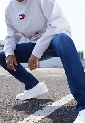 Sudadera gris con un pequeño logo, pantalones vaqueros de mezclilla azul y zapatillas blancas. Manos visibles con anillos, en postura de cuclillas sobre una acera.