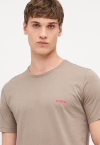 T-shirt à manches courtes couleur brun clair en tissu doux, avec un petit logo rouge "HUGO" sur la zone de la poitrine gauche. Encolure ronde simple.