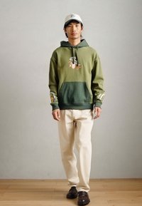 Πράσινη κουκούλα με διαβάθμιση από σκούρο σε ανοιχτό, μπροστινή τσέπη, γραφικό σχέδιο, συνδυασμένη με κρεμ παντελόνι και σκούρα παπούτσια.