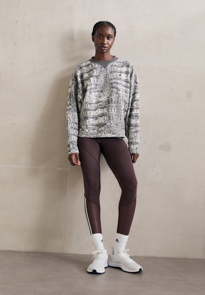 Stående person i en gråmønstret hettegenser, mørkebrune leggings med meshdetaljer, hvite sokker og hvite treningssko foran en ensfarget vegg.