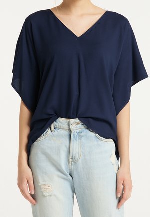 Femme portant un haut bleu marine à col en V avec des manches courtes larges et un jean bleu clair déchiré.