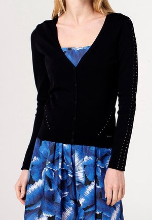 Femme portant un cardigan noir boutonné avec de petits clous sur les manches, par-dessus une robe bleue à fleurs avec de grandes pétales.