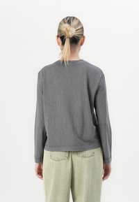 Person with blonde hair in a bun wearing a faded dark gray long-sleeve shirt and light green high-waisted pants, facing away.