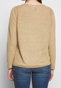 Beige gebreide trui met lange mouwen, ribbelstructuur en een ronde halslijn, gecombineerd met donkerblauwe jeans met zichtbare achterzakken.