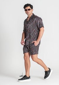 Schwarzes T-Shirt mit kurzen Ärmeln und passende Shorts mit geometrischen Mustern in Pink, Weiß und Gelb. Kombiniert mit schwarzen Turnschuhen und Sonnenbrille.
