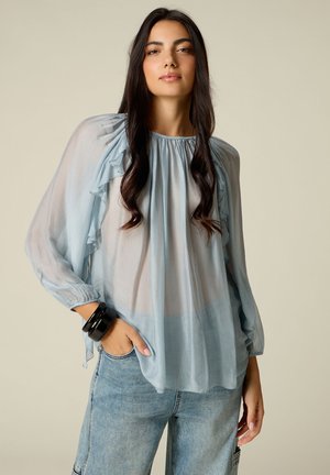 Mujer con cabello largo y oscuro lleva una blusa azul claro transparente con volantes y vaqueros azules claros, posando con una mano en el bolsillo.