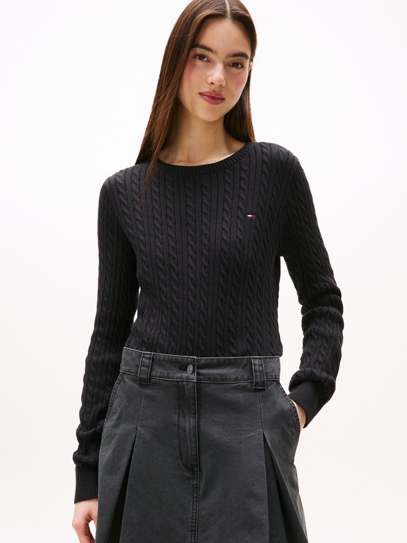 Maglione nero a maglia grossa con scollatura rotonda e polsini a coste, abbinato a una gonna pieghettata grigia con chiusura a bottoni e tasche laterali.