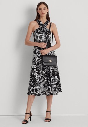 Femme portant une robe midi à fleurs noire et blanche avec un col halter, des sandales à talons noirs et tenant un sac bandoulière noir.