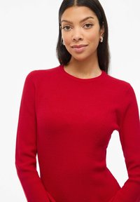 Maglione rosso a coste con maniche lunghe e colletto a girocollo. Presenta un tessuto testurizzato con dettagli di cucitura tonale lungo i lati.