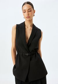 Gilet senza maniche nero con scollatura a V profonda, cintura in vita ben definita e colletto a rever. Il tessuto sembra liscio con un design strutturato e un orlo angolare.