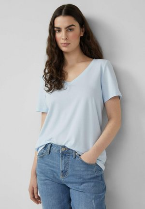 Jonge vrouw met lang krullend haar, gekleed in een lichtblauw T-shirt met V-hals en korte mouwen, en een blauwe spijkerbroek, staand tegen een effen achtergrond.