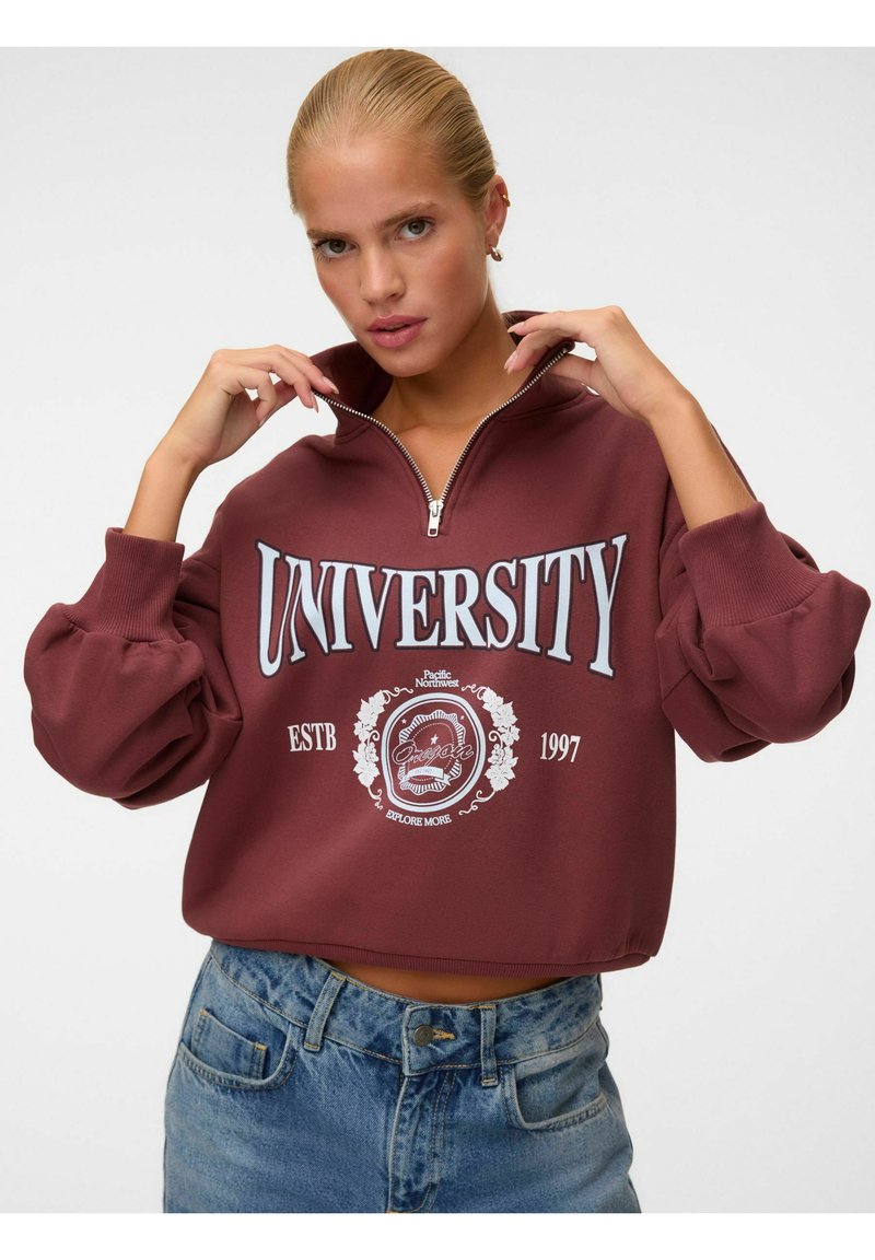 Jeune femme aux cheveux blonds tient le col d'un sweat zippé bordeaux portant l'inscription "University Oregon Pacific Northwest 1997,", habillée d'un jean bleu.