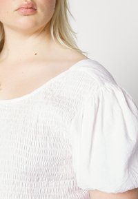 Blusa de algodão branca com textura franzida, decote redondo e mangas curtas bufantes. Tecido suave com uma aparência ligeiramente amarrotada.