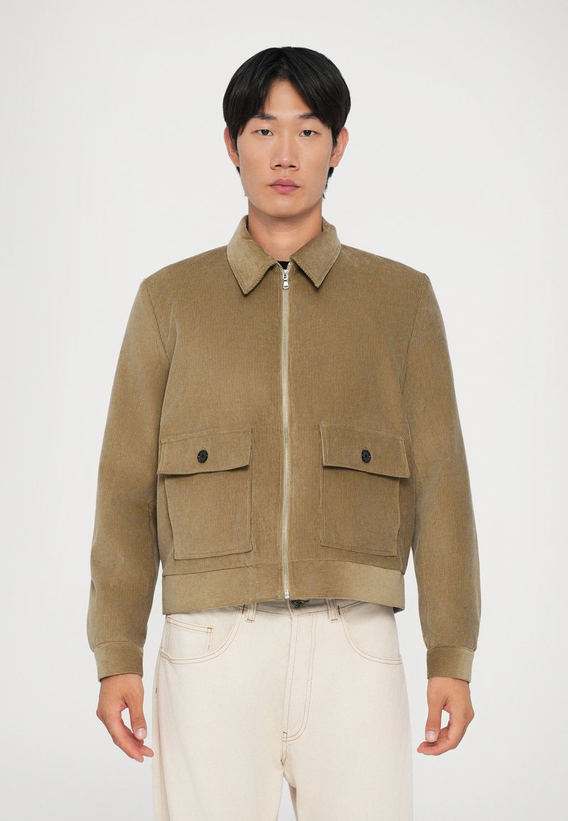 Giacca in corduroy marrone con chiusura a zip, colletto e due tasche sul petto con bottone. Presenta una texture a coste e un design accorciato.