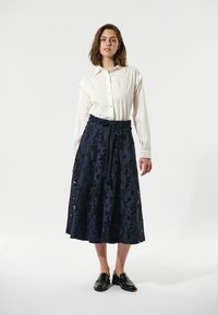 Gonna di pizzo blu marino con motivi floreali, design fluido e vita annodata, abbinata a una camicia bianca con bottoni e mocassini neri.