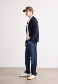 Ναυτικό πλεκτό κασκόλ, λευκό πουκάμισο, μπλε τζιν, λευκά αθλητικά παπούτσια με καφέ σόλες κα�ι καφέ καπέλο, στεκόμενος σε φως φόντο.