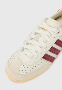 Vit lädersneaker med vinröda mockaränder, perforerad tå och krämfärgade snören; har en texturerad sula och gulddetalj på tungan.