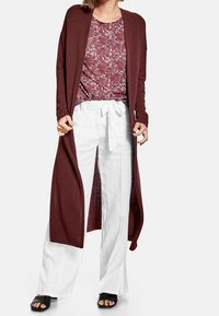 Vinröd cardigan lagd över en mönstrad röd och vit topp, i kombination med vita linnebyxor och svarta halksandaler. Lång, öppen design.