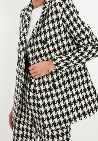 Blazer houndstooth bianco e nero con due bottoni, abbinato a pantaloni coordinati. Un bracciale con catena dorata aggiunge un accento metallico.