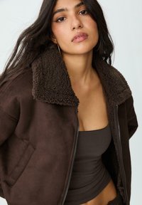 Giacca in suede marrone con colletto in pelliccia shearling morbido, zip frontale, tasche laterali e una texture liscia, indossata sopra un top senza maniche marrone abbinato.