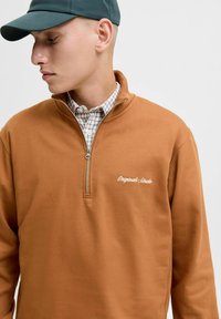 Hnedá zip-up mikina s vyšitým logom, s vysokým golierom a hladkou textúrou, navlečená cez károvanú košeľu.