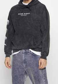 Persona che indossa una felpa con cappuccio nera slavata con scritta "Moon Night" e fasi lunari sulla manica, abbinata a pantaloni di velluto a coste blu scoloriti.