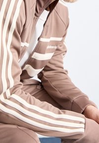 Brun atletisk träningsdräkt med vita randdetaljer och kvartdragkedja. Tillverkad av mjukt material, med en logotyp på bröstet.