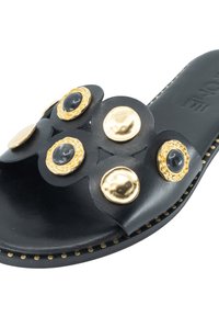 Schwarze Rutsch-Sandale mit strukturiertem Obermaterial und kreisförmigen goldenen und schwarzen Akzenten sowie einer flachen Sohle mit dekorativer Naht entlang des Randes.