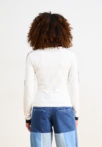 Vit långärmad skjorta med svarta axelränder, kombinerad med blå denimbyxor med blandade tyglappar. Slät textur, avslappnad design.