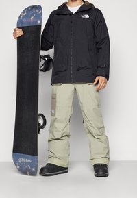 Una persona sostiene una tabla de snowboard negra con acentos en azul y blanco. Lleva una chaqueta impermeable negra y pantalones claros de color beige con bolsillos. Las botas negras completan el look.