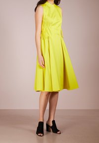 Žluté bezrukávové šaty s přiléhavým korzetem a rozšířenou sukní, se stahovacími detaily v pase. V kombinaci s černými sandály na otevřenou špičku.