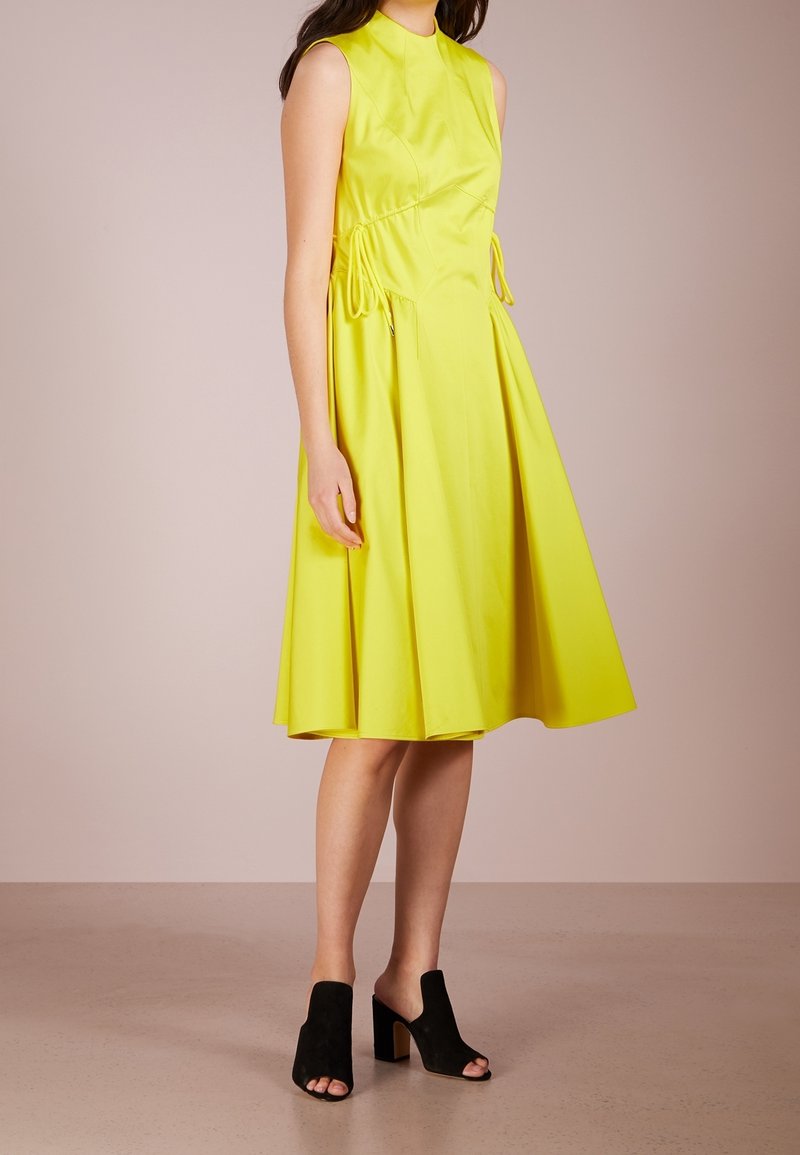 Žluté bezrukávové šaty s přiléhavým korzetem a rozšířenou sukní, se stahovacími detaily v pase. V kombinaci s černými sandály na otevřenou špičku.
