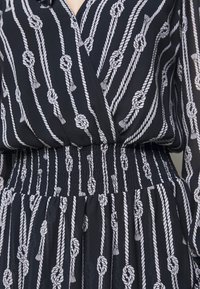 Námořnická blůza s bílým lanovým vzorem, s nařaseným pasem a splývavou tkaninou. Dlouhé, mírně nabírané rukávy s knoflíkovými manžetami.