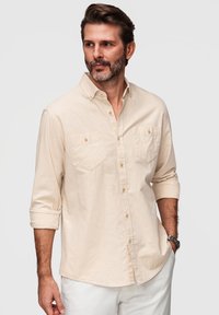 Homme barbu portant une chemise beige à boutons avec les manches retroussées et un pantalon blanc, debout devant un fond clair uni.