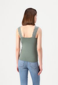Green fitted tank top with lace straps, smooth texture, and a straight hem. Paired with light blue jeans, showing a casual look.