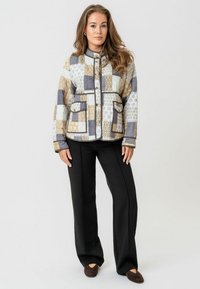 Jachetă matlasată cu un design patchwork în nuanțe de bej, gri și alb, având două buzunare frontale și o închidere cu nasturi. Asortată cu pantaloni negri.