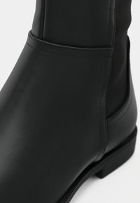 Bottine en cuir lisse noir avec coutures visibles et petit talon, présentant des détails latéraux et avant.