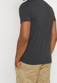 Camiseta de color gris oscuro, de manga corta, hecha de una tela suave. Tiene un cuello redondo y un pequeño detalle de logo en la manga. Combinada con pantalones de color beige claro.