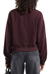 Persoon met krullend haar, gekleed in een textuur donkerpaarse trui en vervaagde zwarte jeans, met een kleine zwarte handtas aan de zijkant.