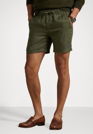 Olivengrønne lærredsshorts med elastisk talje og snøre, med en blå logoaccent på siden. Bæres med brune loafers.