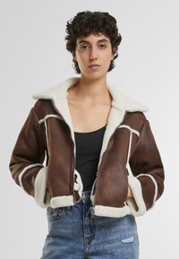 Giacca corta in ecopelle marrone con foderatura in shearling bianco, colletto ampio e cuciture a contrasto. Chiusura con zip e tasche laterali.