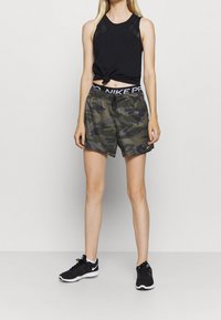 Top deportivo negro sin mangas con un detalle anudado, combinado con pantalones cortos de camuflaje verde con una cintura elástica. Zapatillas negras completan el conjunto.