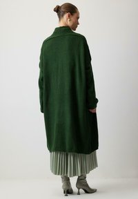 Long green cardigan with a relaxed fit, made of soft material, featuring dropped shoulders. Worn over a pleated light green skirt and gray boots.