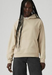 Sudadera beige de tela suave con un bolsillo frontal, capucha con cordón y puños acanalados, combinada con pantalones gris oscuro. Diseño simple y casual.