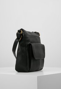Svart läder crossbody-väska med framficka och justerbar rem, uppställd på en vit pelare mot en enkel grå bakgrund.