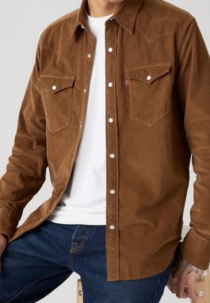 Mężczyzna ubrany w brązową koszulę sztruksową na guziki z przednimi kieszeniami na klapki, na białej koszulce i niebieskich dżinsach, siedzący na drewnianym stołku.