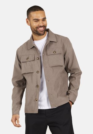 Lächelnder Mann, der eine taupefarbene, geknöpfte Jacke mit zwei Brusttaschen über einem weißen Hemd und einer schwarzen Hose trägt, und mit einer Hand in der Tasche posiert.